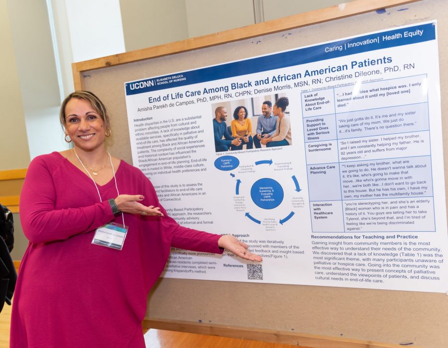 A presenter stands beside her research poster titled “End of Life Care Among Black and African American Patients,” gesturing toward the display.