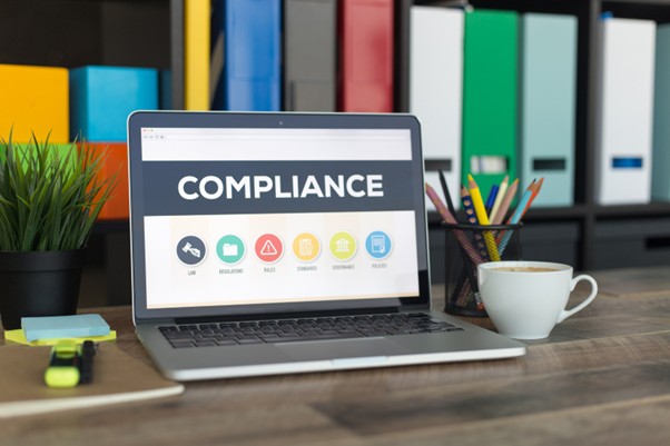 A laptop displaying the word "Compliance" on the screen. On the desk beside it are a cup of coffee, sticky notes, and a highlighter.