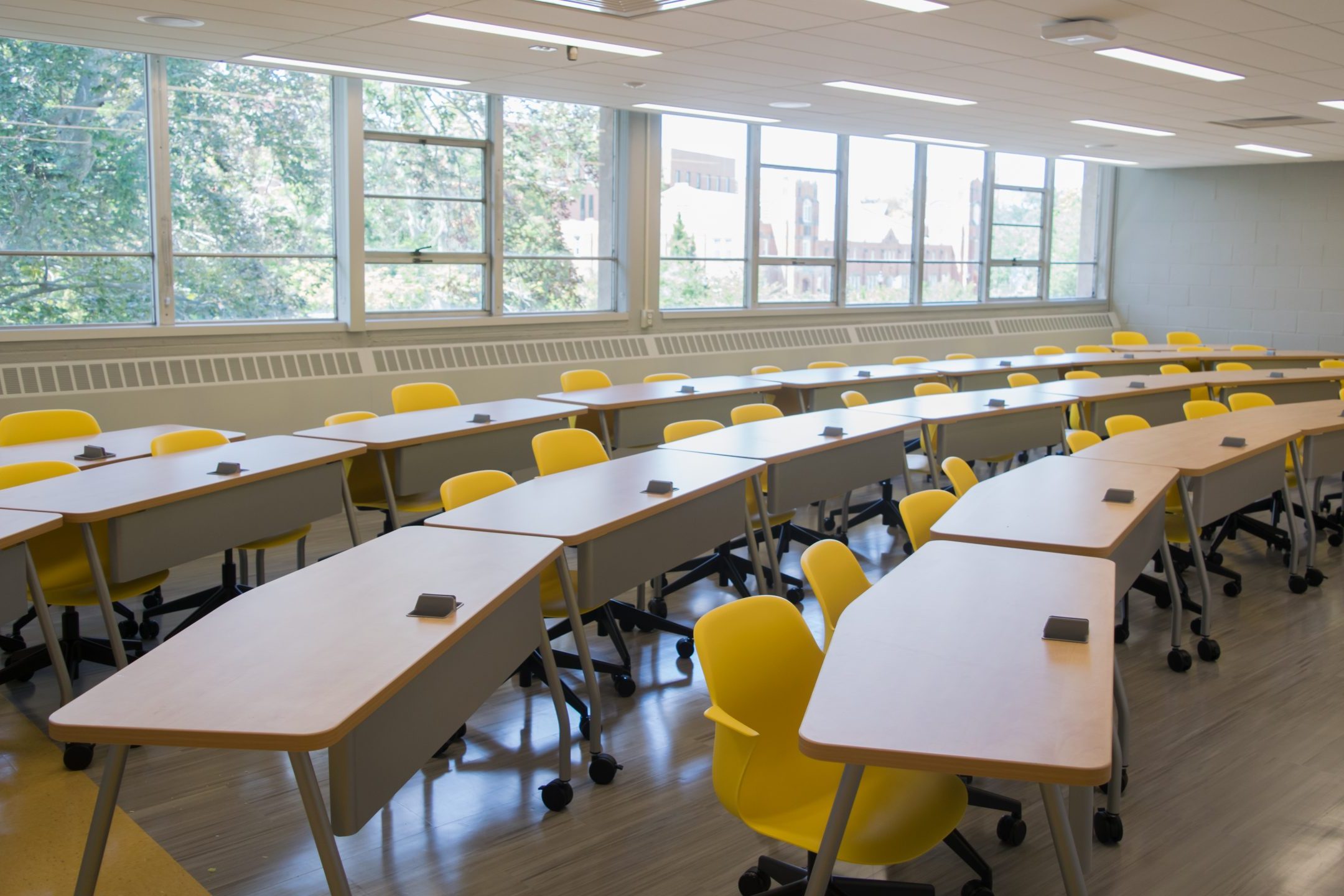 Classroom with desks, yellow chairs, and a wide window showing green foliage.