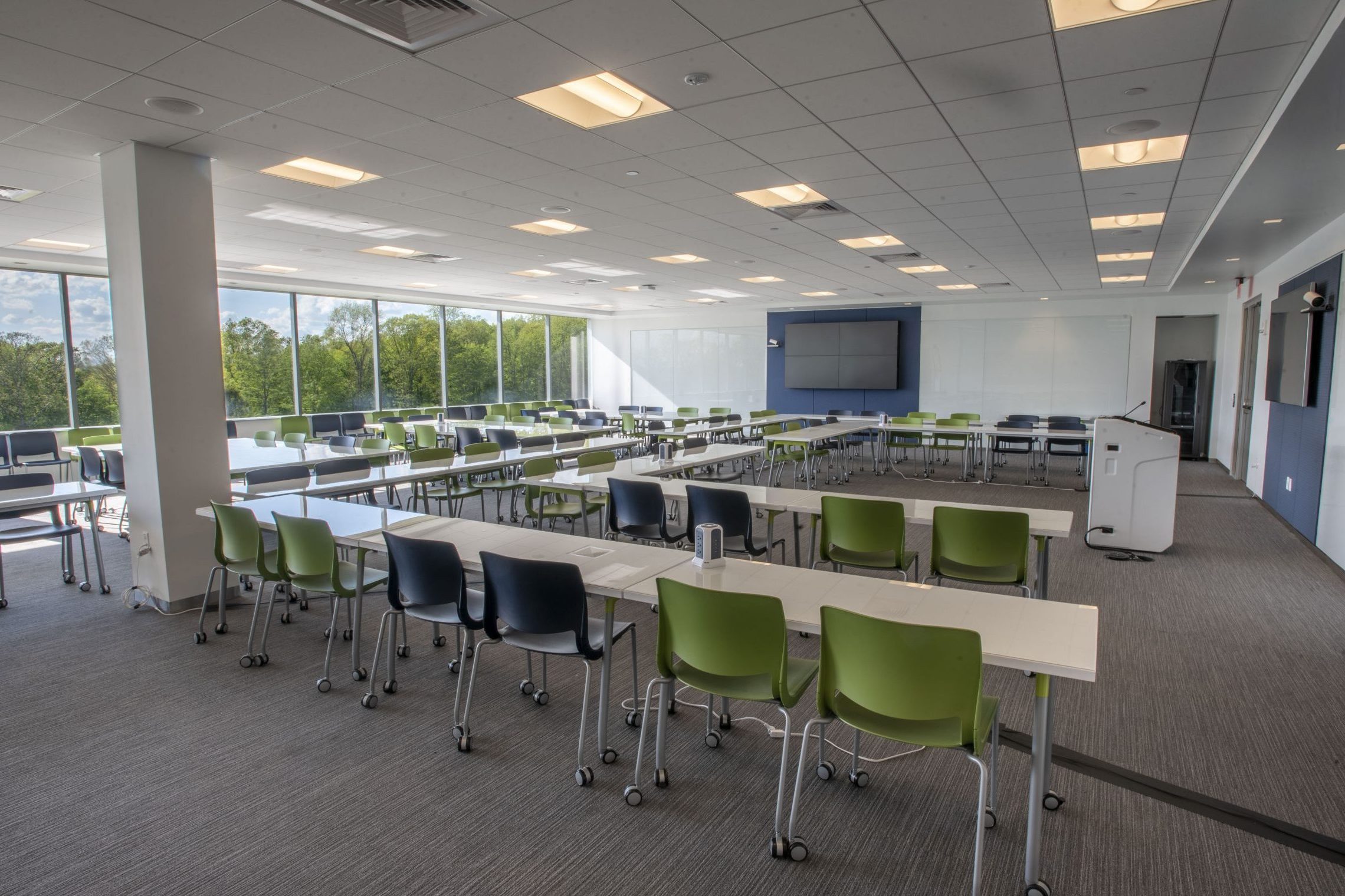 A classroom with several tables and chairs arranged in a horseshoe-like pattern, two television screens mounted on the walls, a podium, and a window revealing green trees outside.